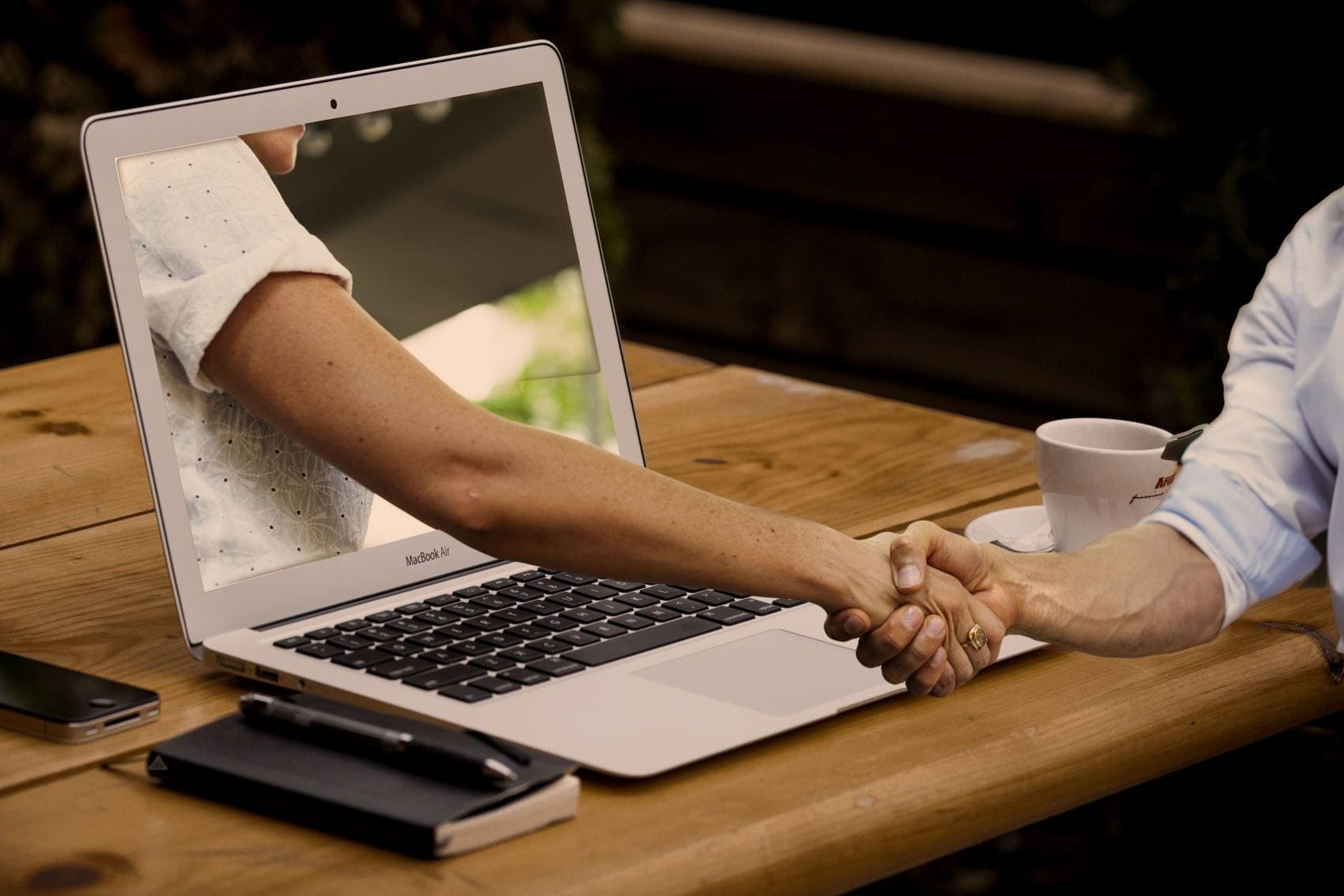 Two people shaking hands.  One person is on one side of a laptop computer, and the other person appears to be inside the screen of the computer, with their hand reaching through the screen to connect with the hand of the other person.