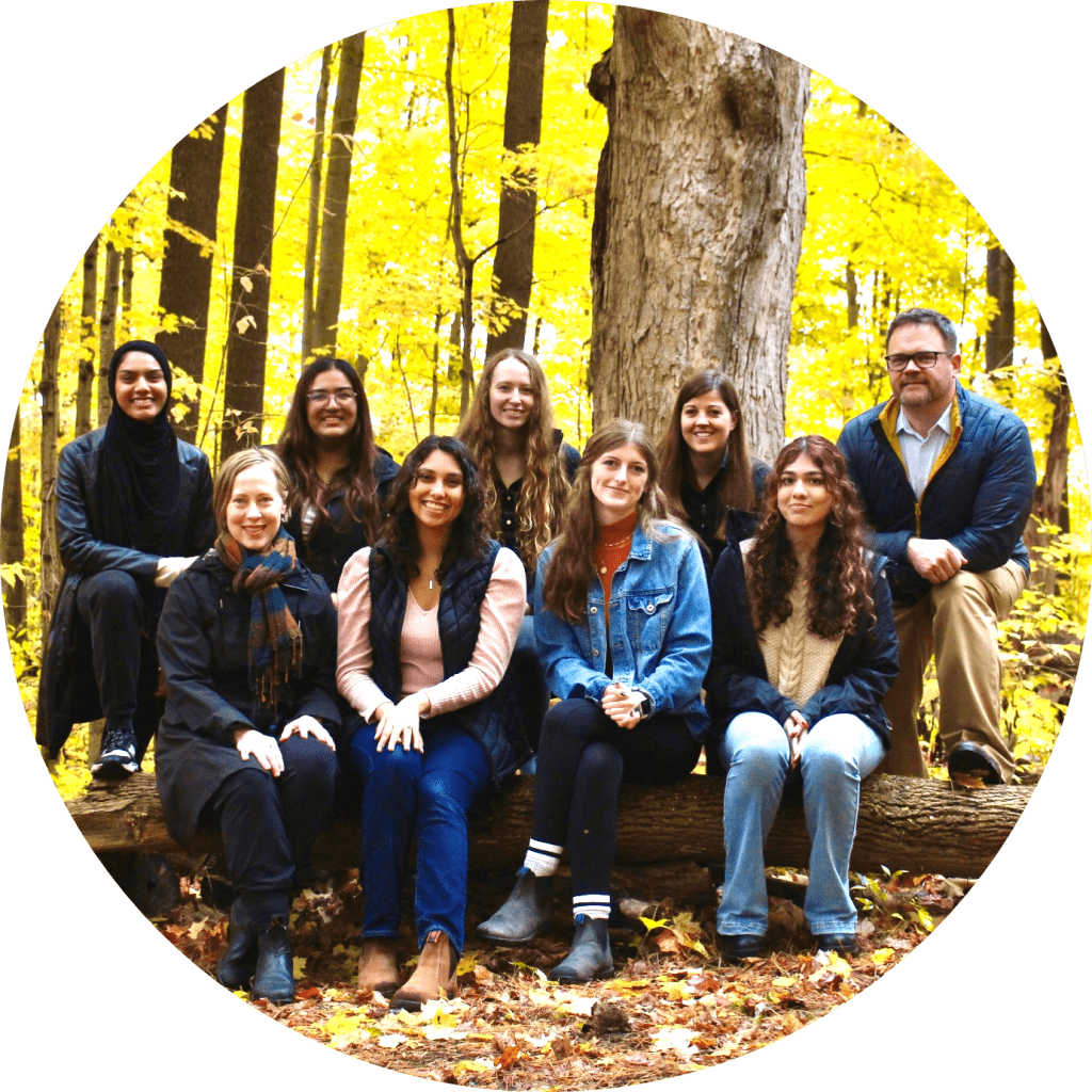 RCVM@OVC team posing for a group portrait, sitting on a log in a wooded area of the Arboretum at the University of Guelph.